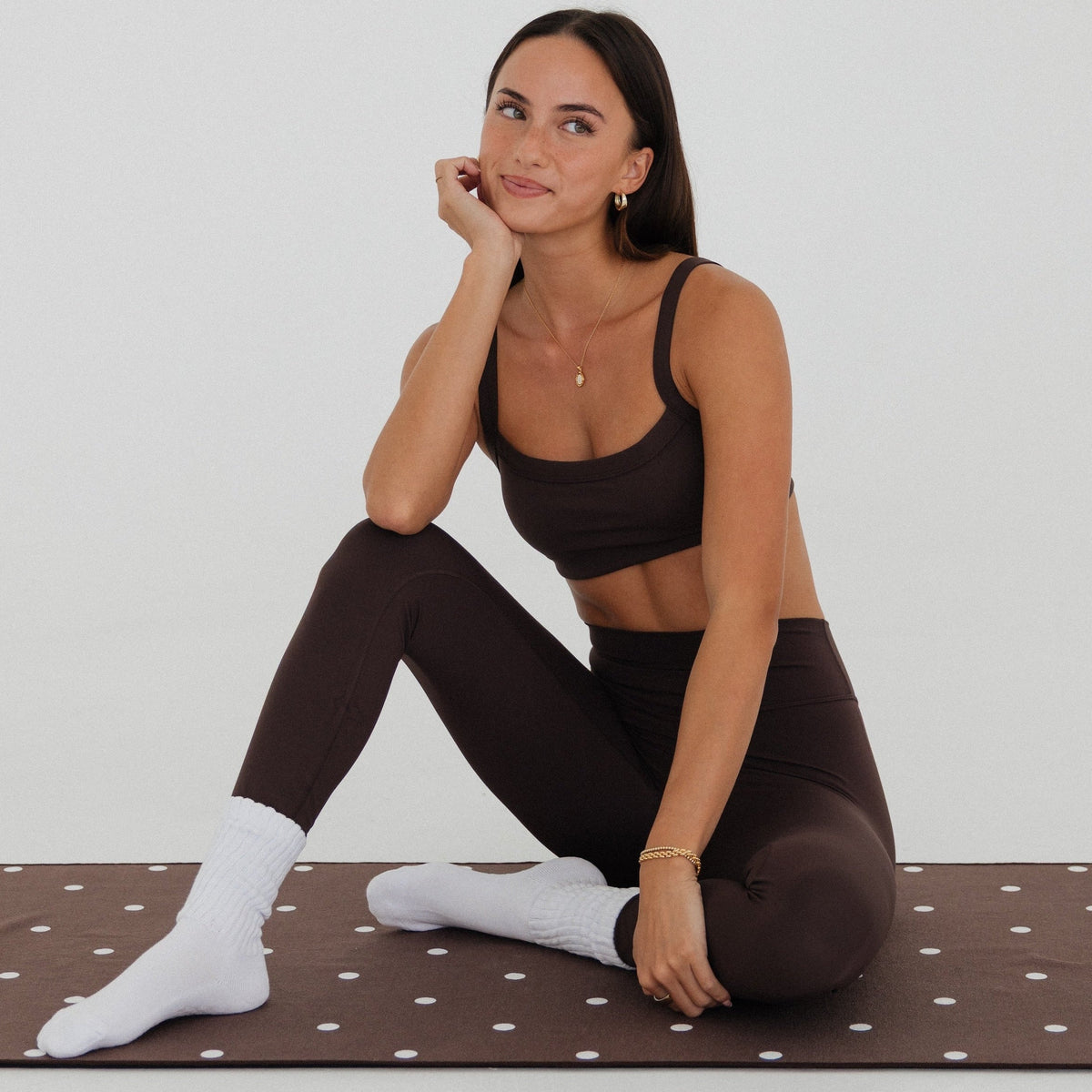 A woman sits on a brown yoga mat with white polka dots, wearing a matching brown sports bra and leggings set with white socks, against a plain light gray studio background.