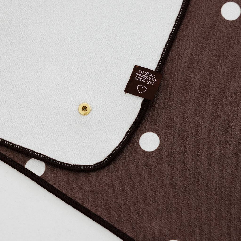 Dark brown yoga mat towel with white polka dots and gold buttons. Towel folded to show white back with tag that states "o Small With Great Love"