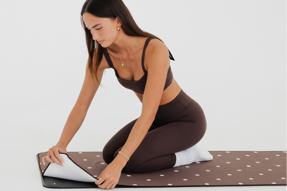 Woman in brown athletic wear rolling out a yoga mat on a white background